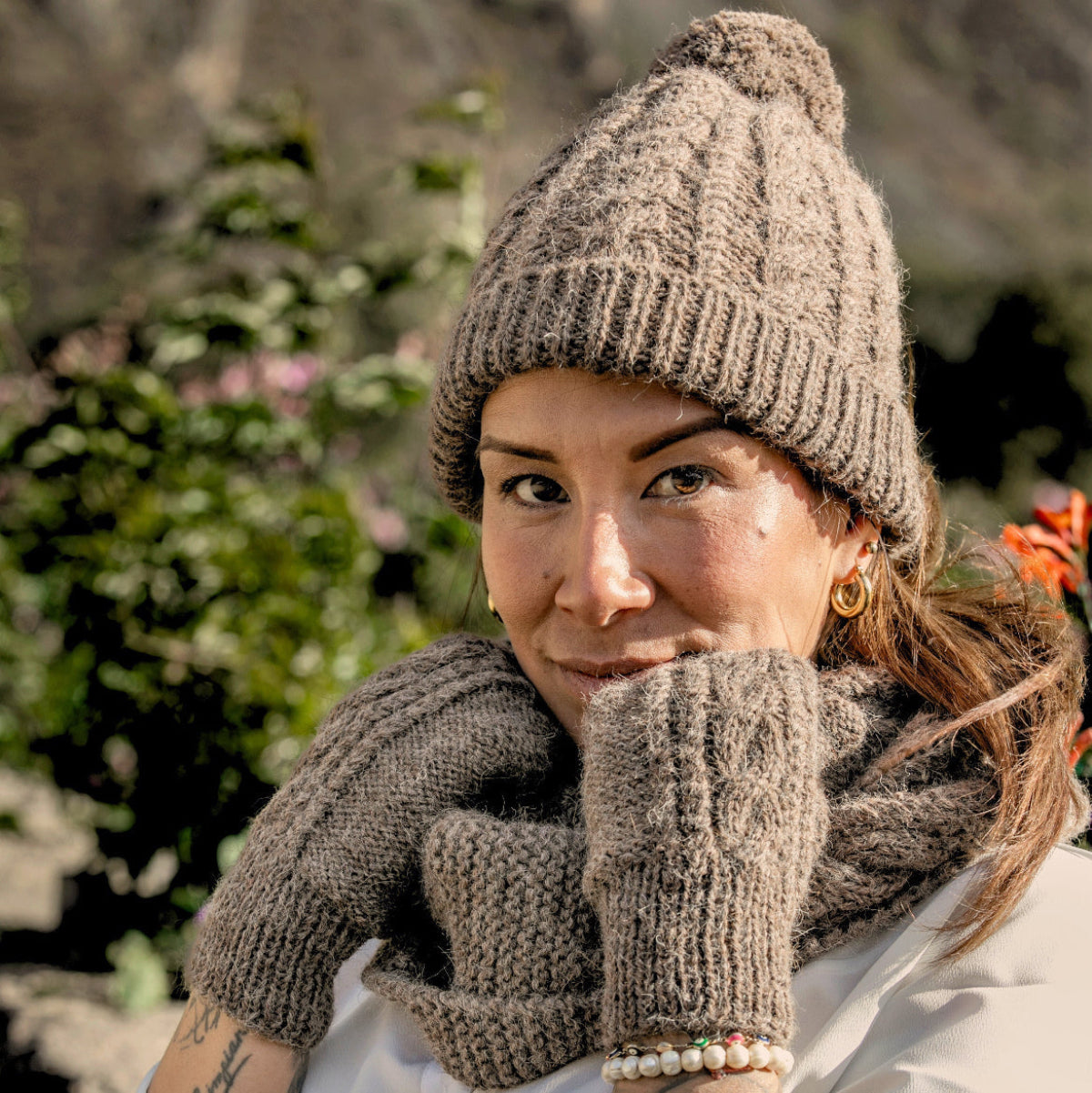 A woman wearing a chunky knit beanie, matching knit mittens, and a knit cowl scarf in a taupe brown color, posing outdoors .