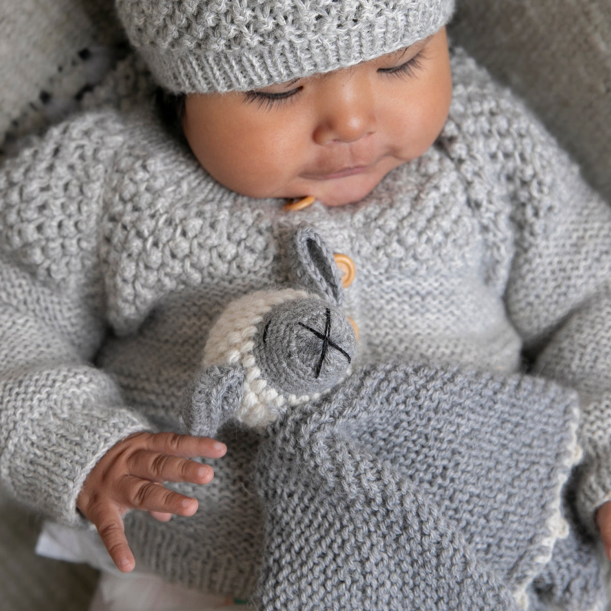 Baby in a gray knit outfit holding a plush toy on a textured surface