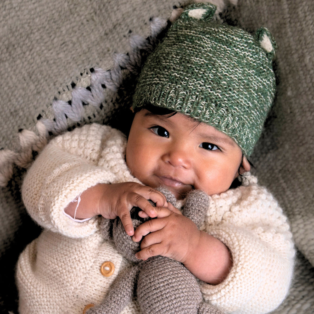 A baby lying on a textured gray blanket, wearing a cream-colored knit cardigan and a green knit hat with small white accents.