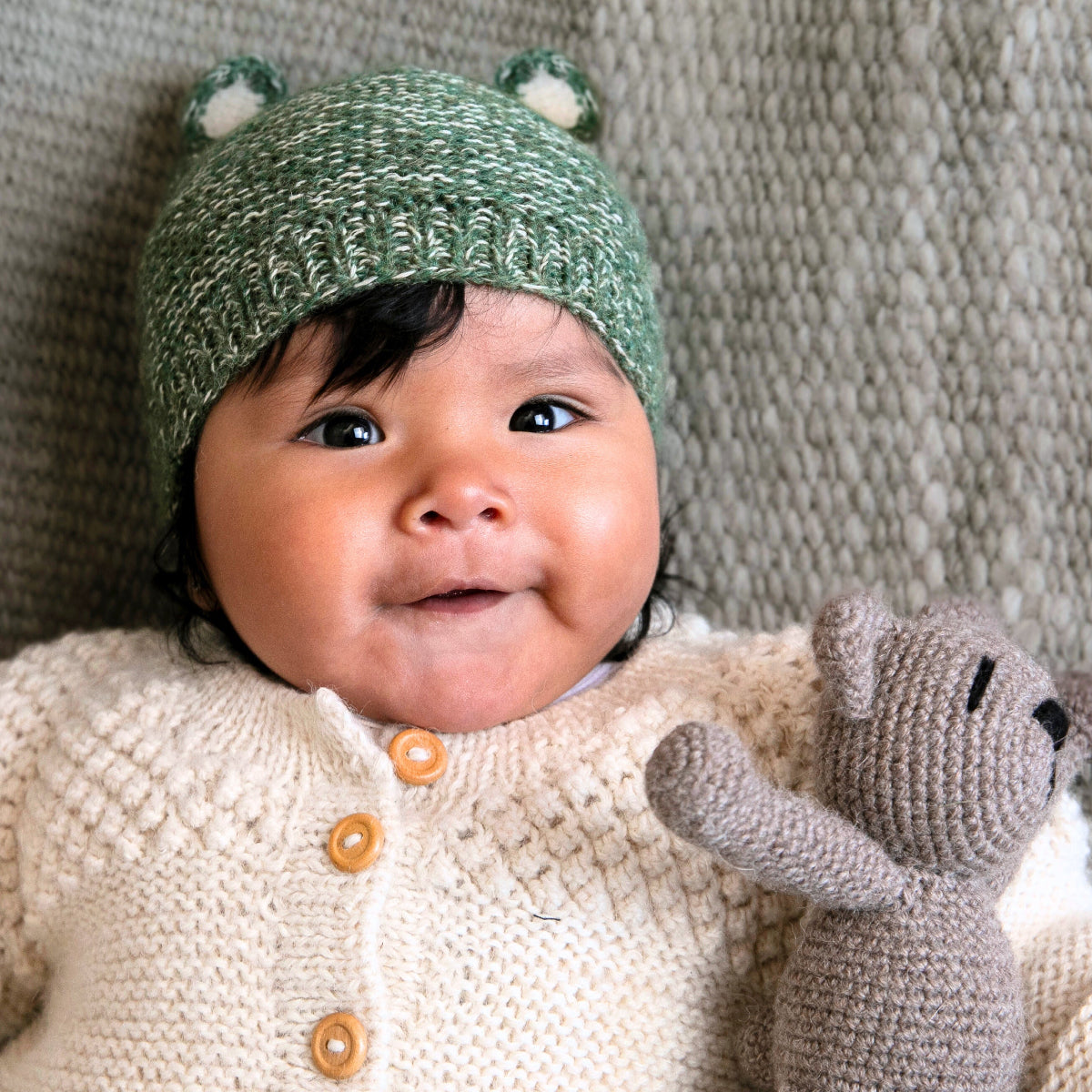 A baby lying on a textured knit blanket, wearing a green crocheted hat with small ear details and a cream knit cardigan, while holding a gray stuffed animal.