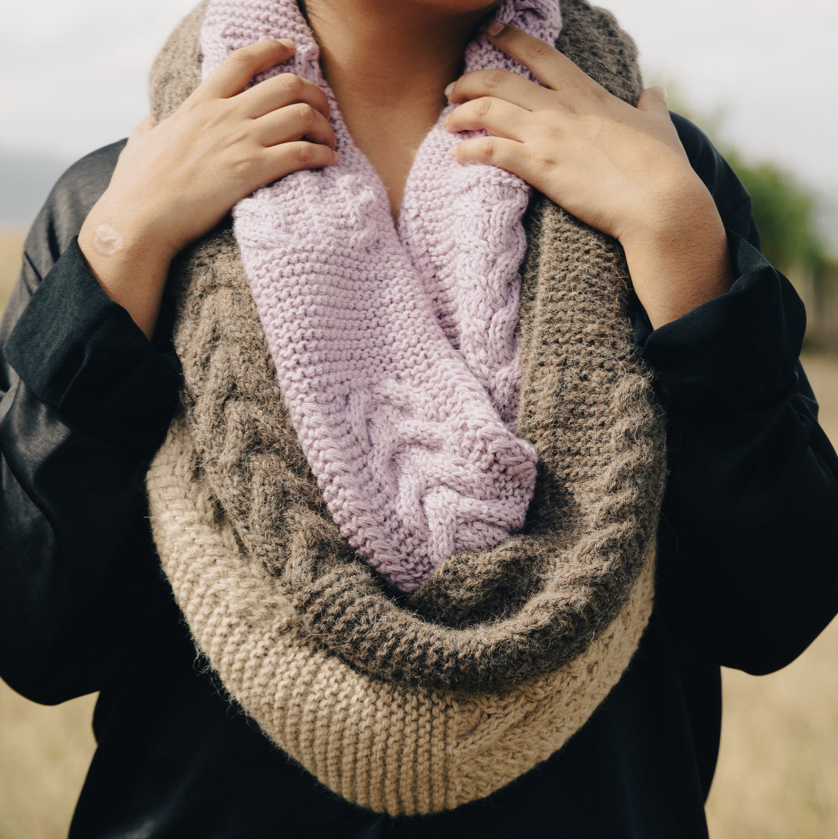 A person outdoors wearing a thick cable-knit infinity scarf in a two-tone design of taupe brown and light pink.
