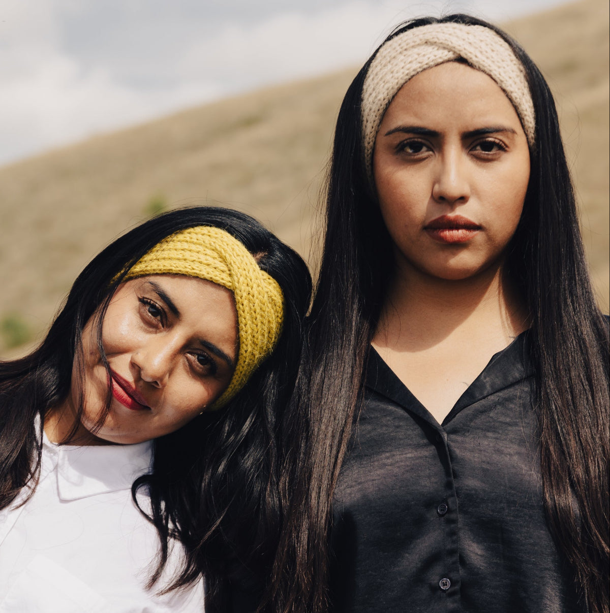 Two young women posing outdoors against a dry grassy hill under an overcast sky. The woman on the left wears a white button-up shirt and a yellow knit headband, while the woman on the right wears a black long-sleeve button-up shirt, a beige knit headband, and brown pants.