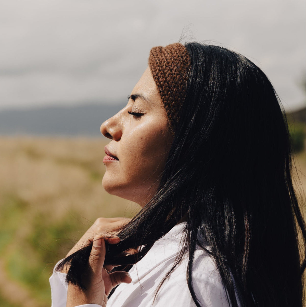 A woman in profile wearing a light pink knit headband and a black satin jacket, set against a blurred outdoor background of mountains and a cloudy sky.