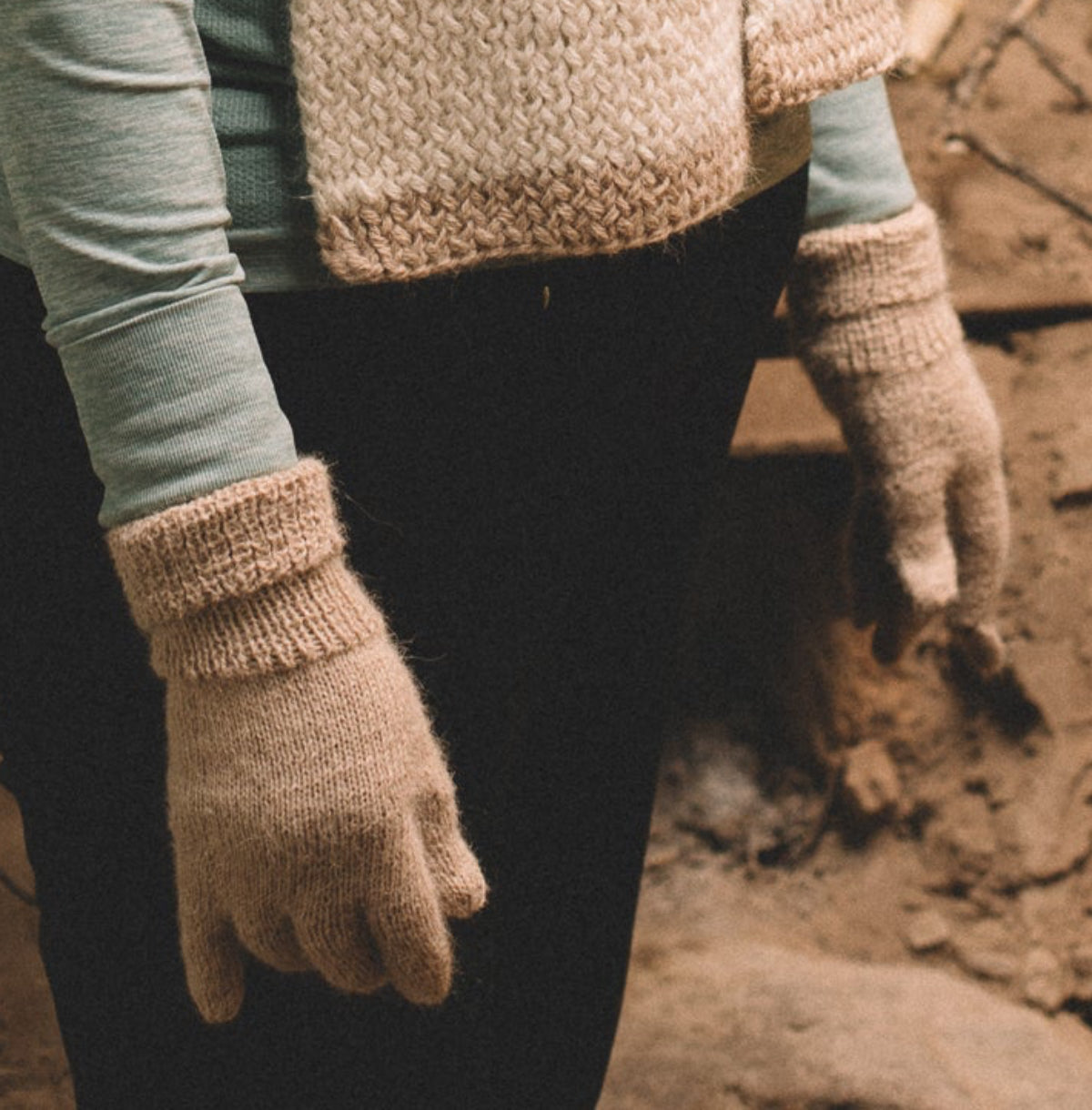 A woman wearing a chunky knit cream scarf, a light blue long-sleeve shirt, and cream knit gloves stands in a rustic, earthy room with a clay stove in the background.