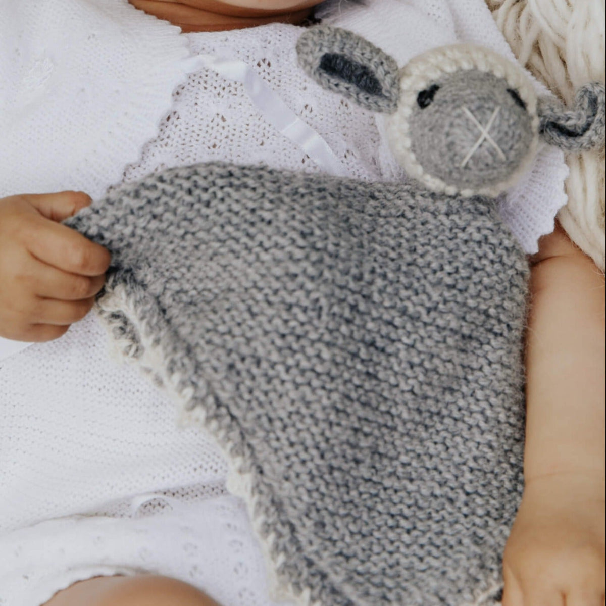 A close-up of a baby wearing a white knit romper while holding a gray knitted security blanket with a sheep head.