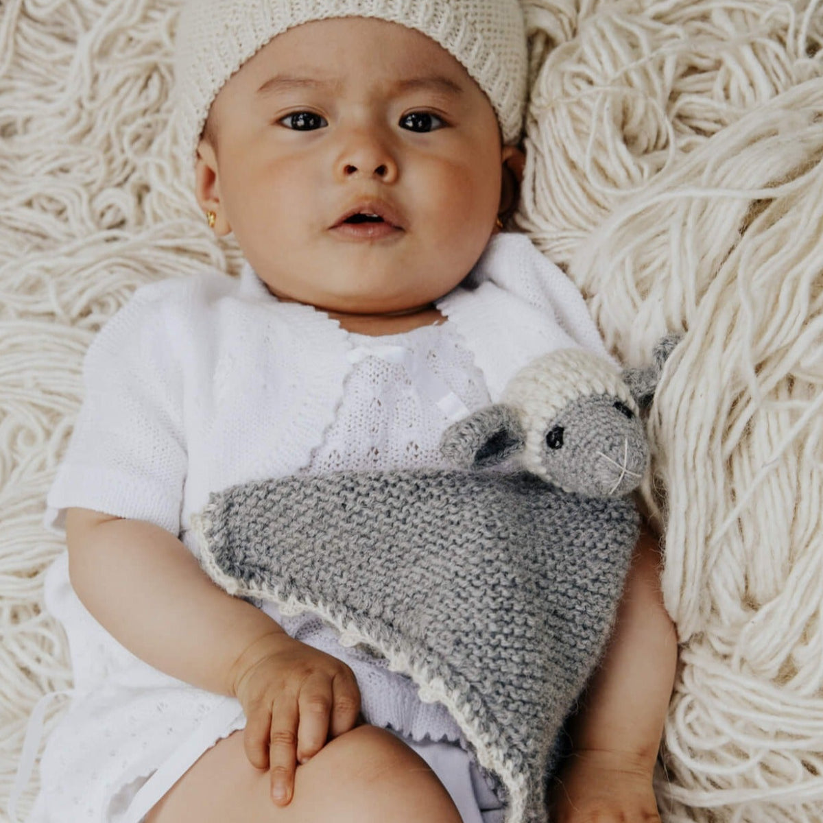 A baby lying on a cream-colored yarn blanket, wearing a cream knit beanie and white outfit, while holding a gray knitted security blanket with a sheep.