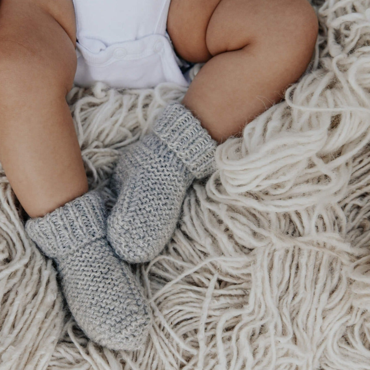 A close-up of a baby's legs wearing light gray knit booties or bootie socks, sitting on a thick, cream-colored shaggy blanket.