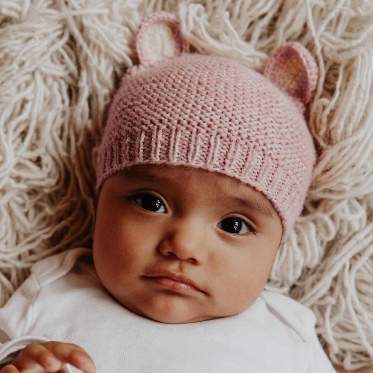 A baby wearing a light pink knit hat with small ear details, lying on a soft cream-colored textured blanket.