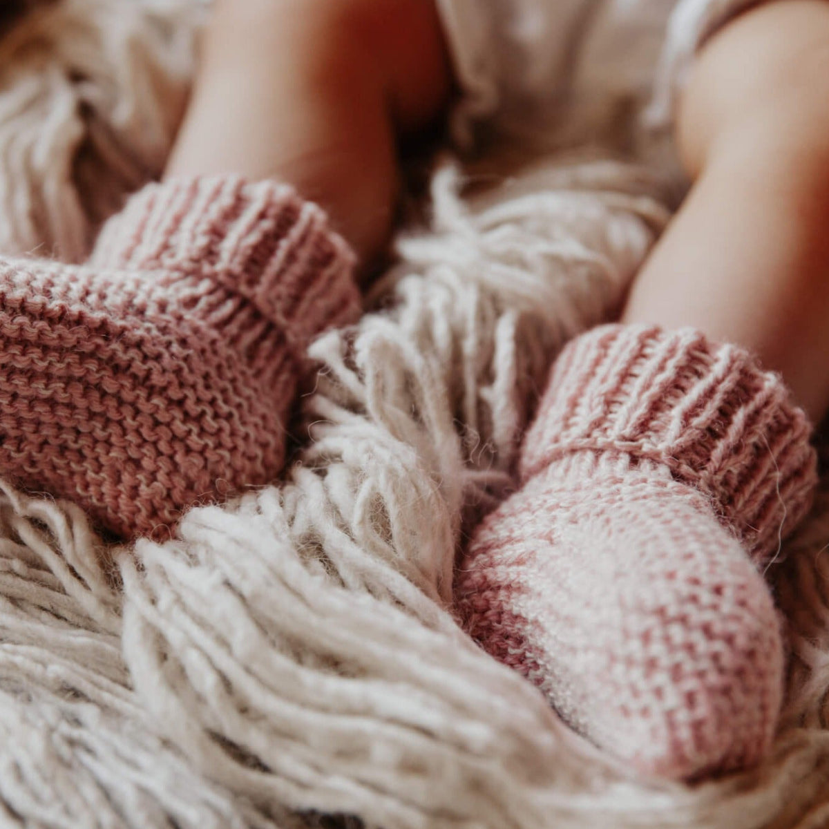 A close-up of a baby's legs wearing dusty pink hand-knit booties resting on a fluffy cream-colored blanket.