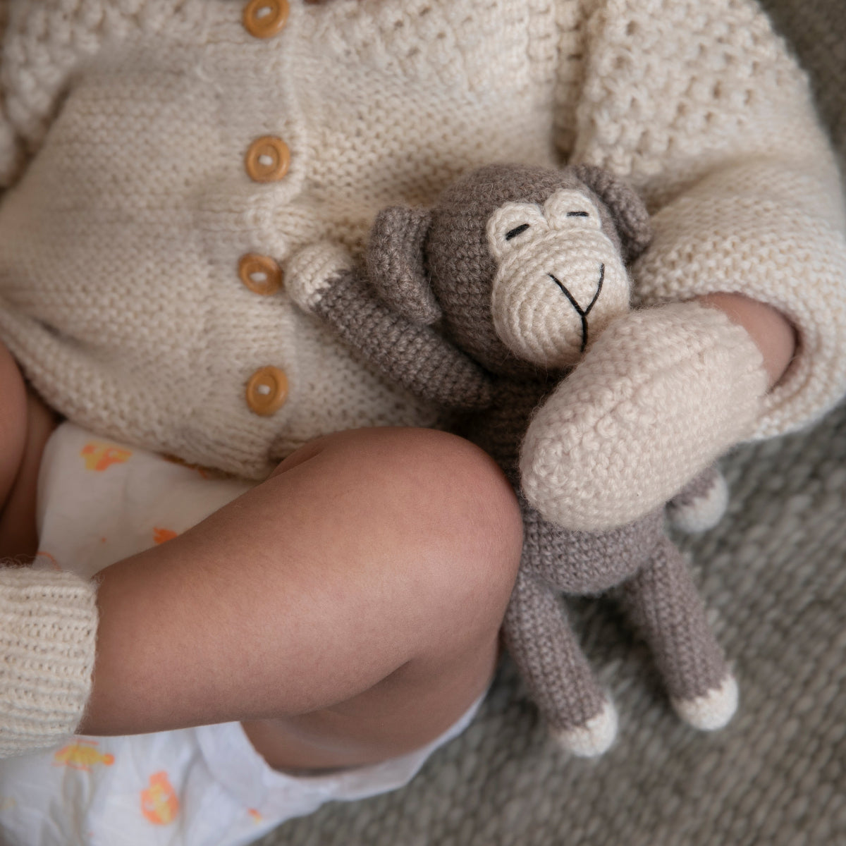 A close-up of a baby wearing a cream knit cardigan and matching knit booties, holding a small gray plush monkey toy.