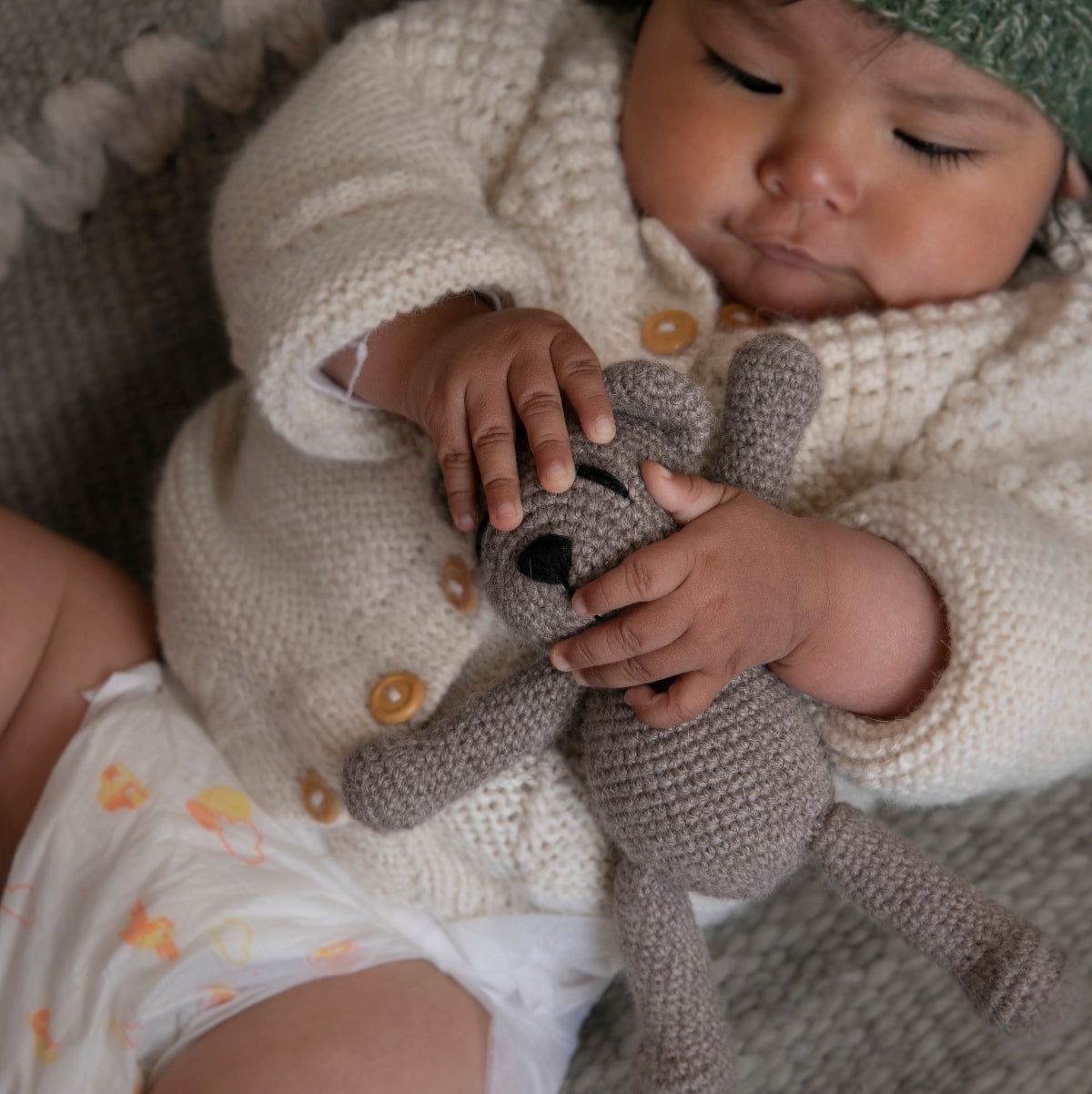 A baby lying on a textured gray blanket, wearing a cream knit cardigan, white diaper, and green knit hat, while holding a small gray crocheted stuffed bear.